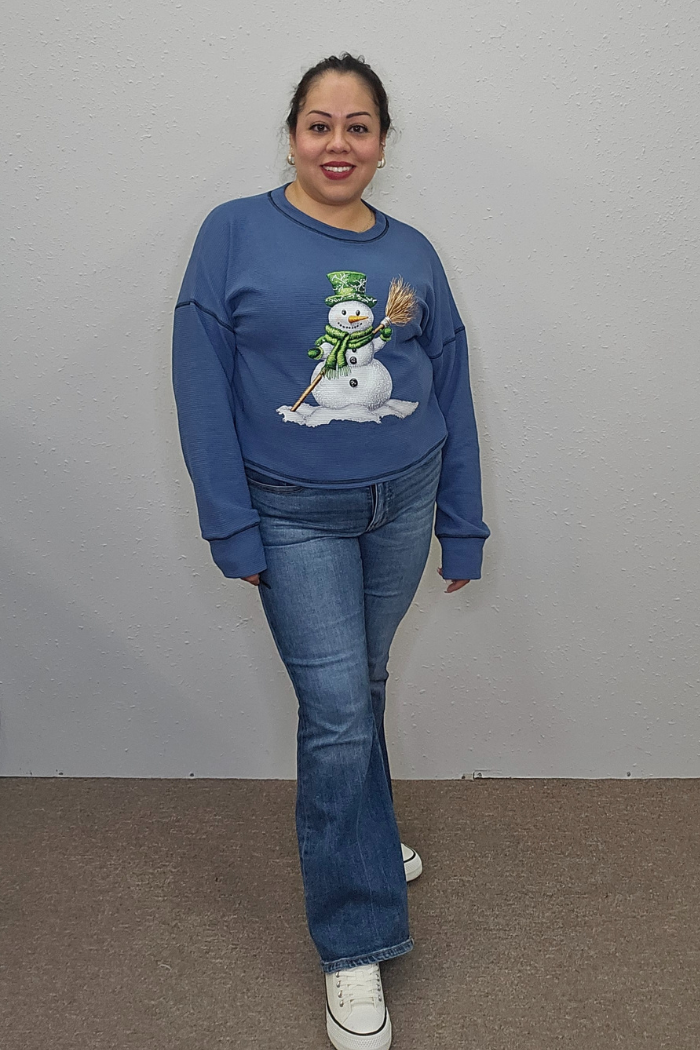 A woman smiles against a light wall, wearing the Snowman Holiday Waffle Top with festive embroidery, paired with blue jeans and white sneakers.