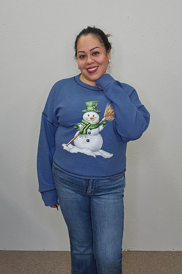 A smiling woman stands against a plain light wall, wearing the Snowman Holiday Waffle Top featuring charming holiday embroidery and blue jeans. Her hair is tied back, and she rests one hand on her neck.
