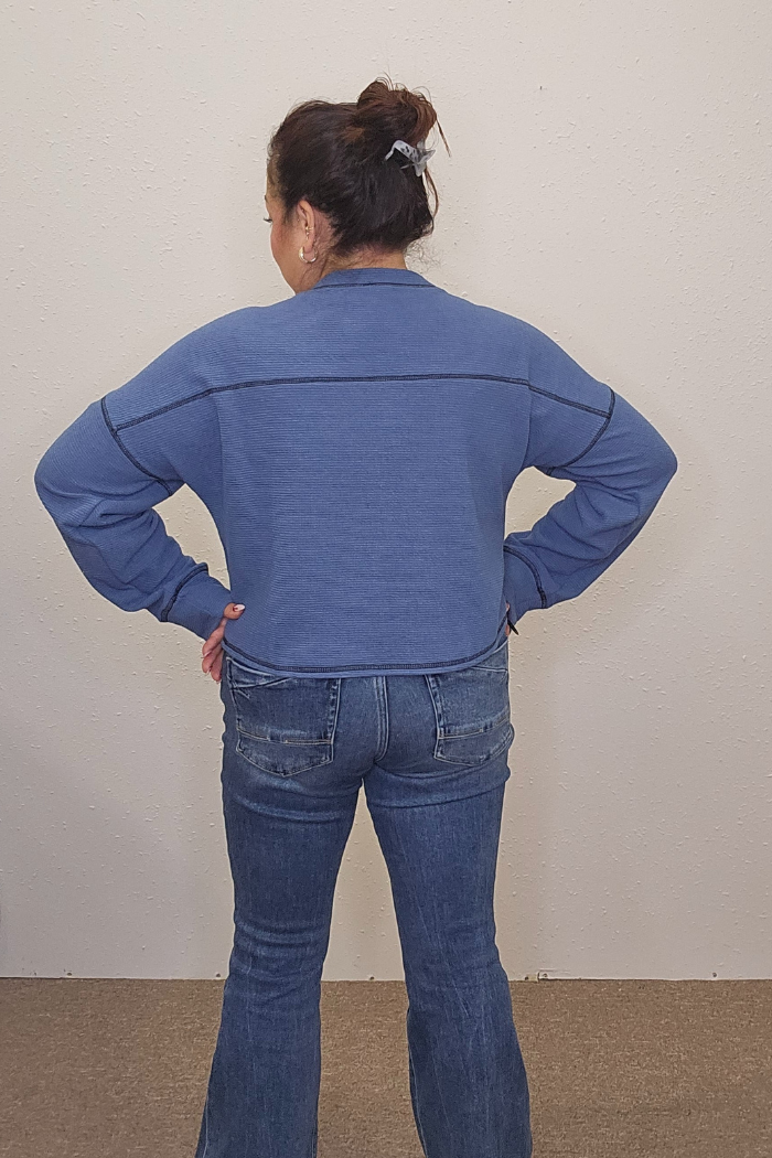 A person with dark hair in a bun, wearing a Snowman Holiday Waffle Top and blue jeans, stands facing a beige wall with hands on hips.