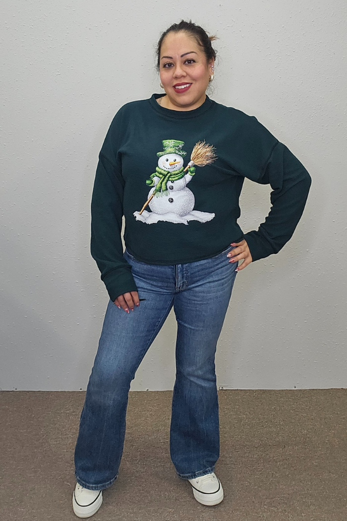 A smiling woman stands against a plain wall, wearing the dark green Snowman Holiday Waffle Top with holiday embroidery, paired with blue jeans and white sneakers. Her hair is tied back and one hand rests on her hip.