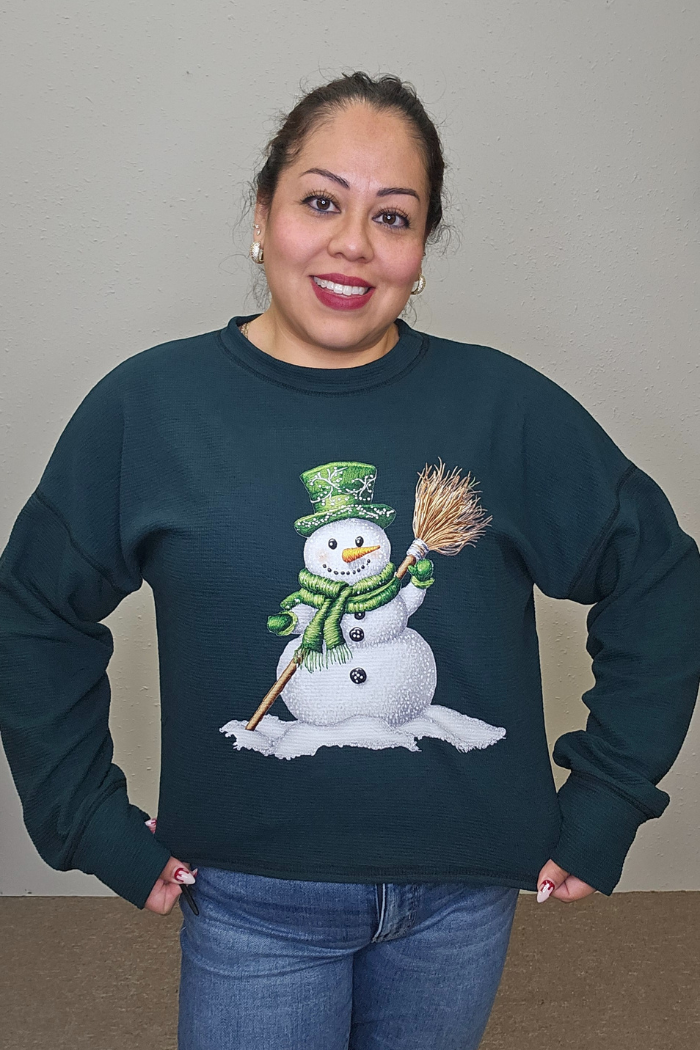 A woman smiles at the camera, wearing the Snowman Holiday Waffle Top—a dark green sweater with holiday embroidery featuring a snowman in a green hat, scarf, and mittens. She stands against a plain beige wall with her hands on her hips.
