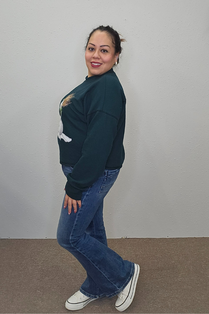 A woman with a ponytail smiles at the camera in a dark green Snowman Holiday Waffle Top, blue jeans, and white sneakers, posing sideways against a plain light wall.