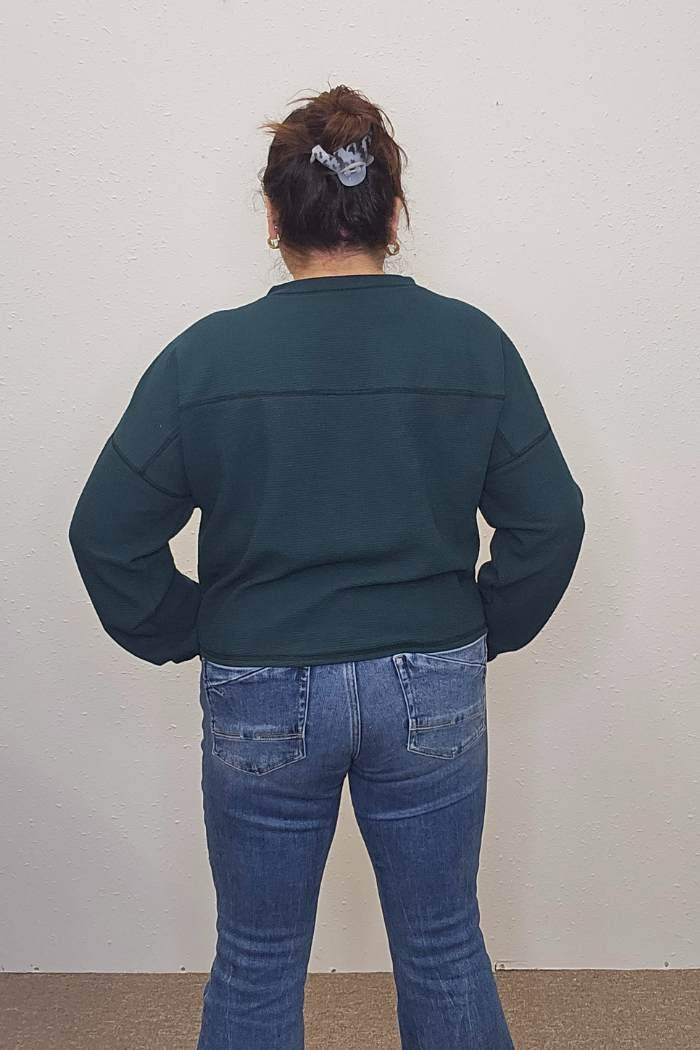 A person with dark hair in a scrunchie, wearing the Snowman Holiday Waffle Top and blue jeans, stands with their back to the camera facing a plain light wall.