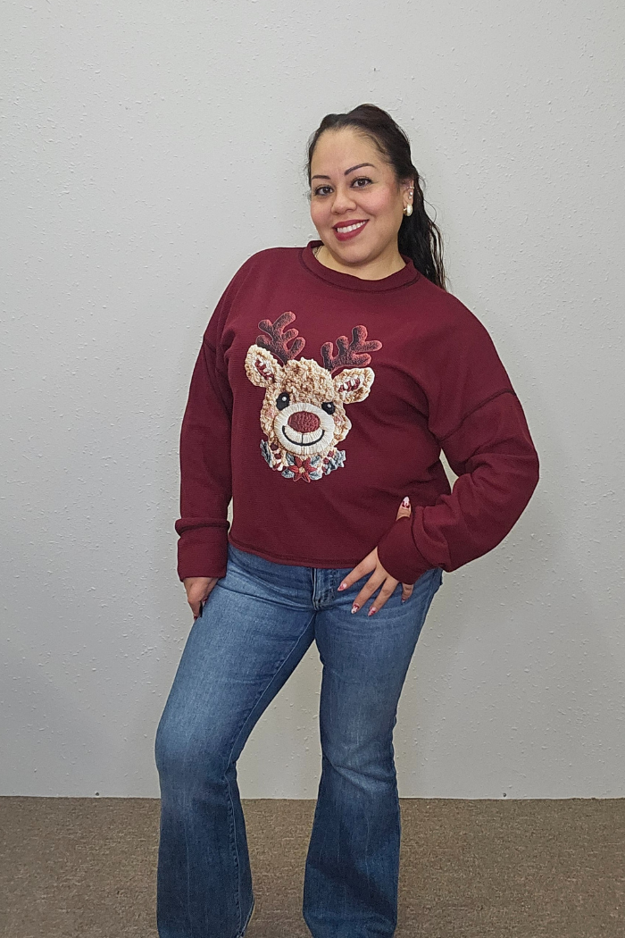 A woman smiles against a plain wall, wearing the Rudolph Holiday Waffle Top with a sequined reindeer design and blue jeans.