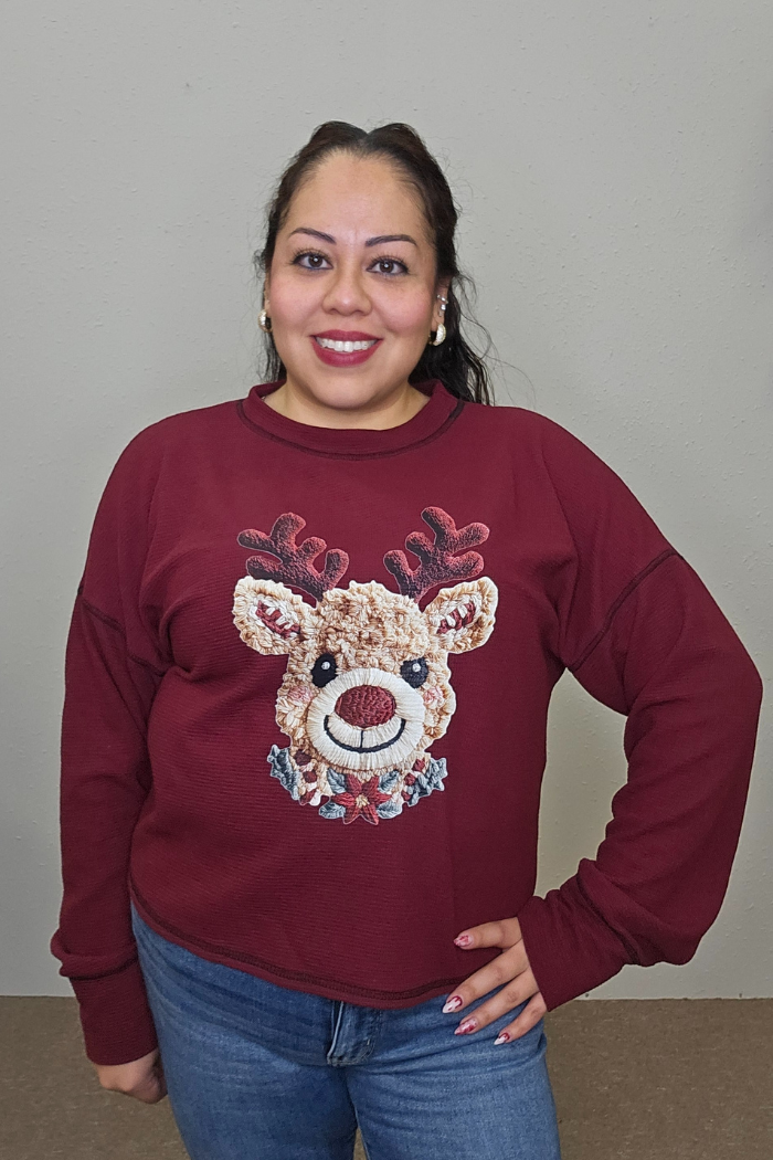 A woman with long dark hair smiles at the camera, wearing the Rudolph Holiday Waffle Top and blue jeans. She stands in front of a plain beige wall with her hand on her hip.
