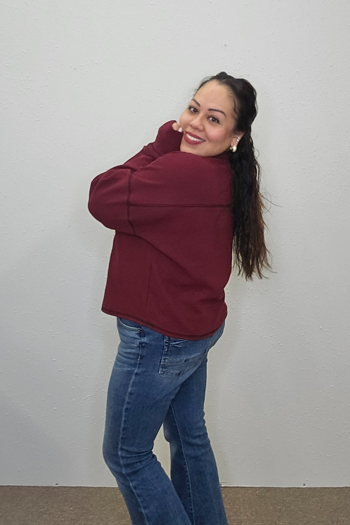 A woman with long dark hair wears the Rudolph Holiday Waffle Top and blue jeans, standing sideways and smiling at the camera in front of a plain light-colored wall.