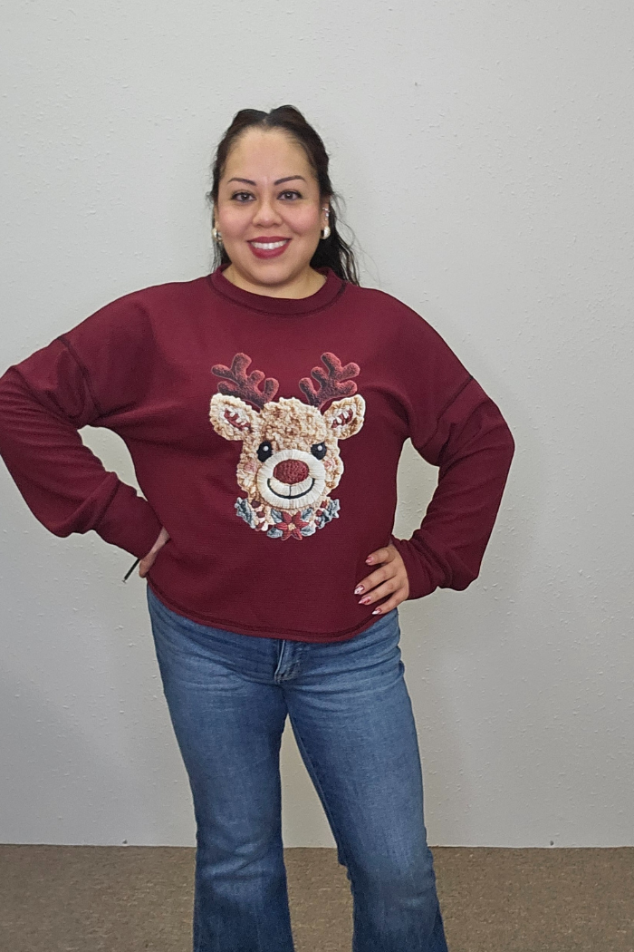 A woman smiles with her hands on her hips, wearing the Rudolph Holiday Waffle Top—a cozy oversized sweatshirt with a cartoon reindeer—paired with blue jeans, standing in front of a plain light-colored wall.