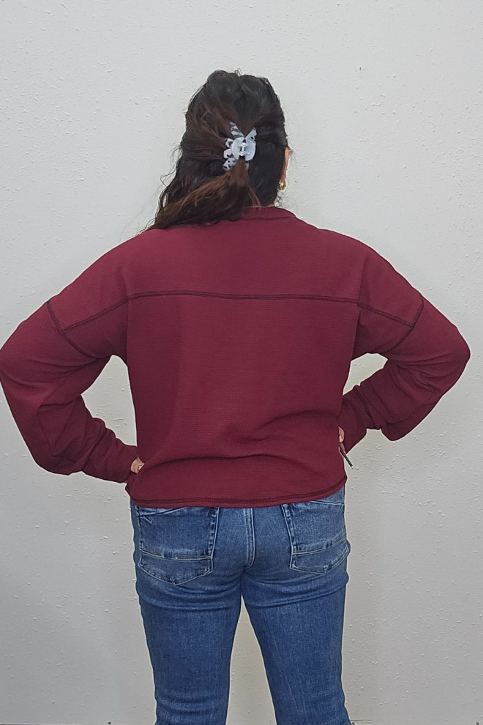 A person stands facing a white wall with hands on hips, wearing the Rudolph Holiday Waffle Top, blue jeans, and a gray scrunchie in their hair. Their back is visible.