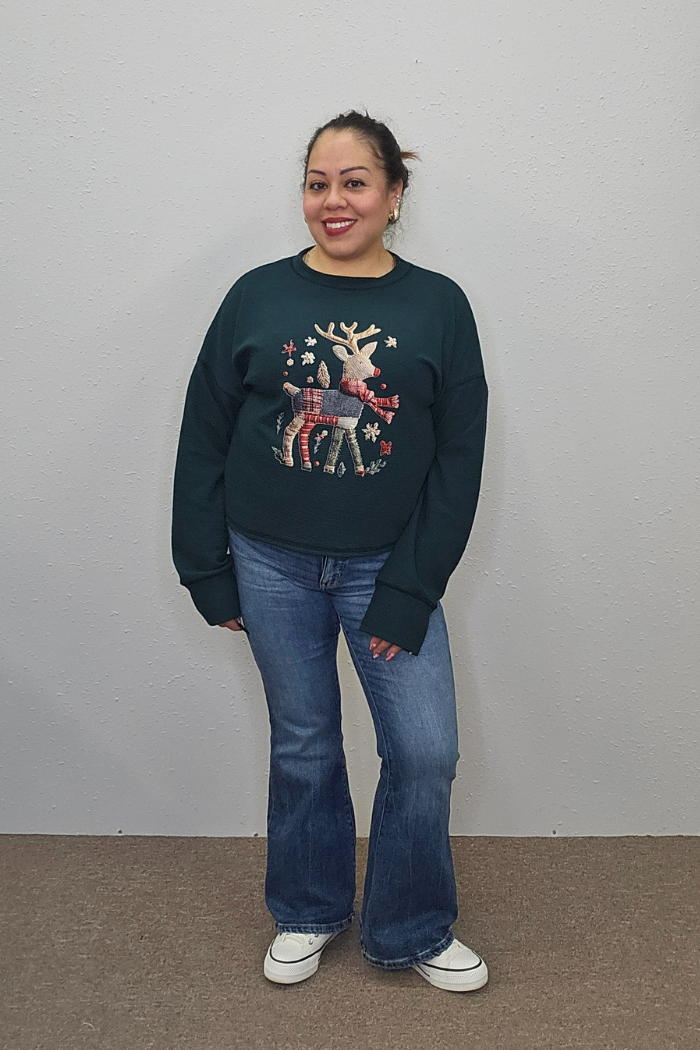 A woman smiles against a plain light wall, wearing the Reindeer Holiday Waffle Top with a festive design. She pairs it with blue jeans and white sneakers.