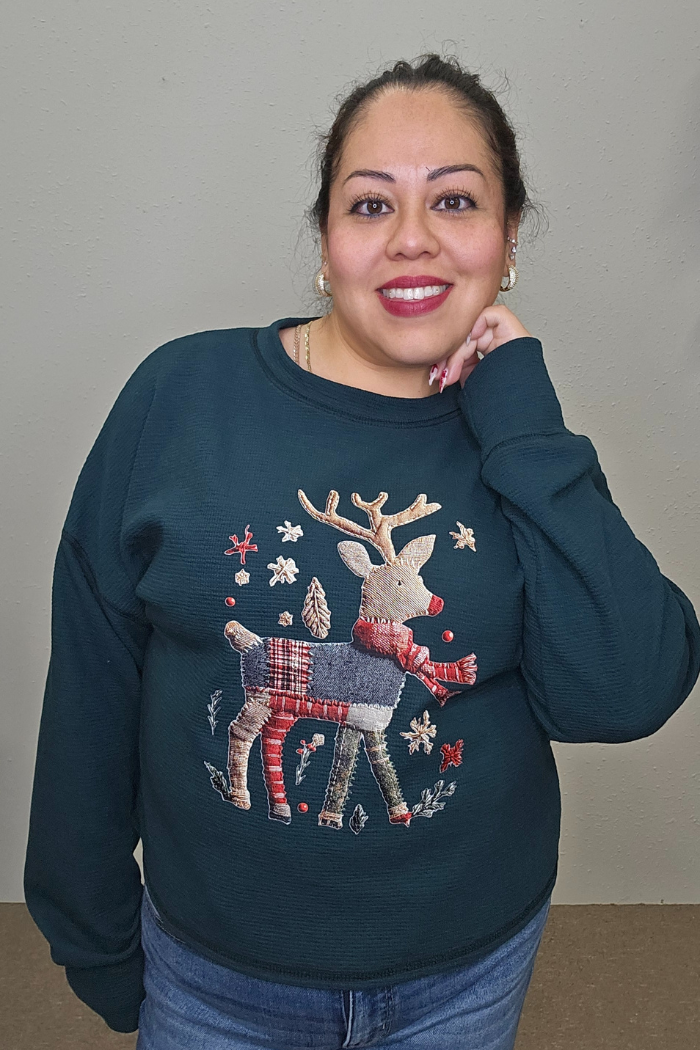 A woman with dark hair tied back smiles while wearing the oversized Reindeer Holiday Waffle Top, featuring patchwork and festive details. She poses with one hand on her face against a plain background.