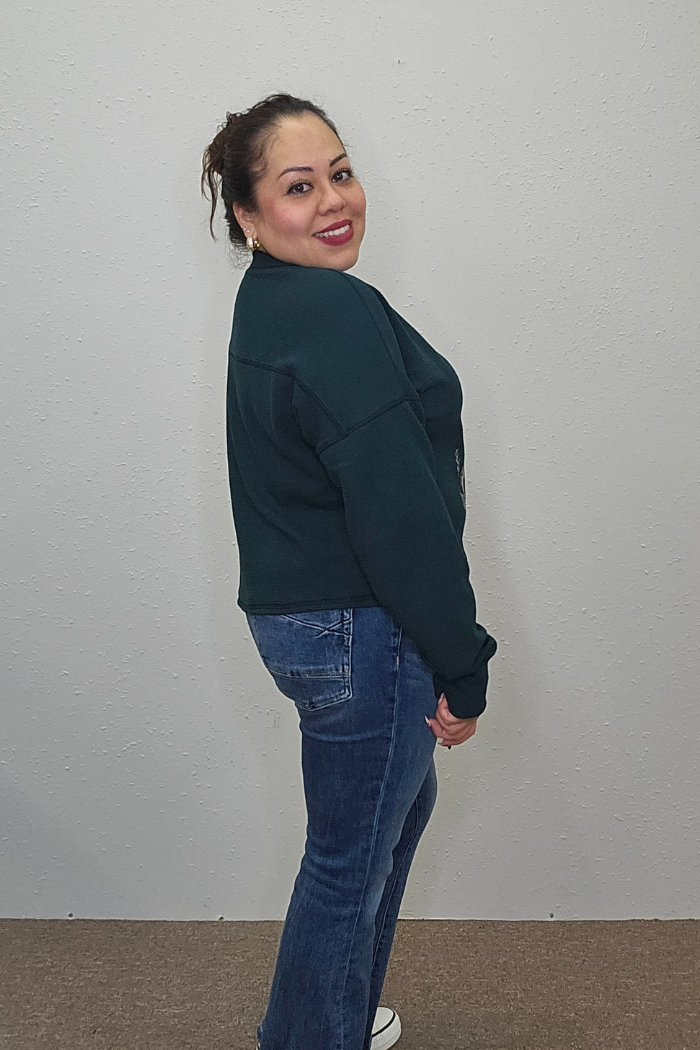 A woman with dark hair in a ponytail, wearing the Reindeer Holiday Waffle Top, blue jeans, and white shoes, stands sideways and smiles at the camera in front of a plain light-colored wall.