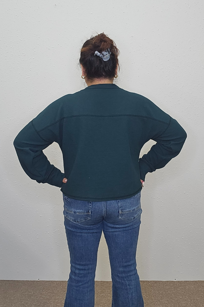A person with dark hair in a scrunchie stands facing a white wall, wearing the Reindeer Holiday Waffle Top and blue jeans, with hands resting on their hips.