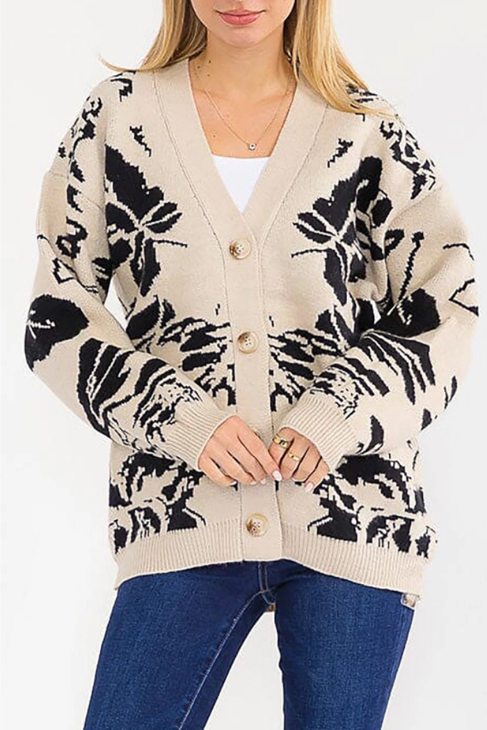 A woman wears the Multi Print Button Cardigan, featuring black floral patterns and front buttons, over a white top with blue jeans. She stands facing forward, hands clasped together.