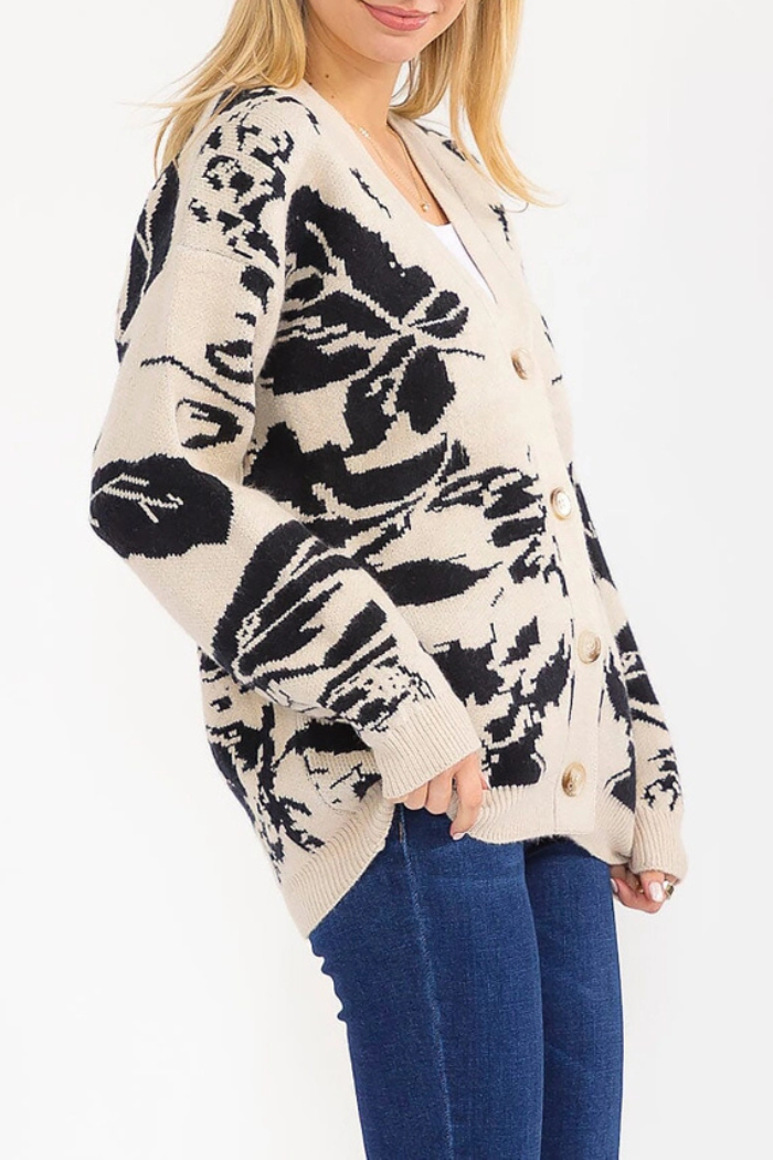 A woman with blonde hair models the Multi Print Button Cardigan, featuring black abstract floral patterns and front buttons, paired with blue jeans against a white background.