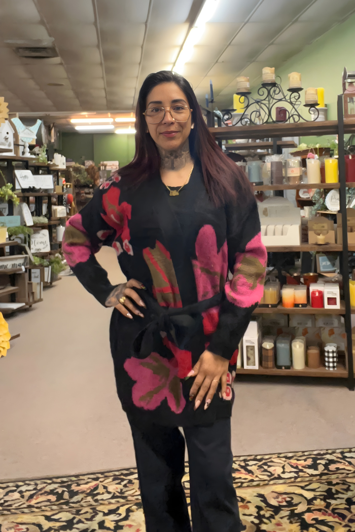 A woman with long, dark hair and glasses stands on a patterned rug inside a home decor store, smiling with one hand on her hip. She wears the Floral Belted Knit Cardigan adorned with large pink flowers.
