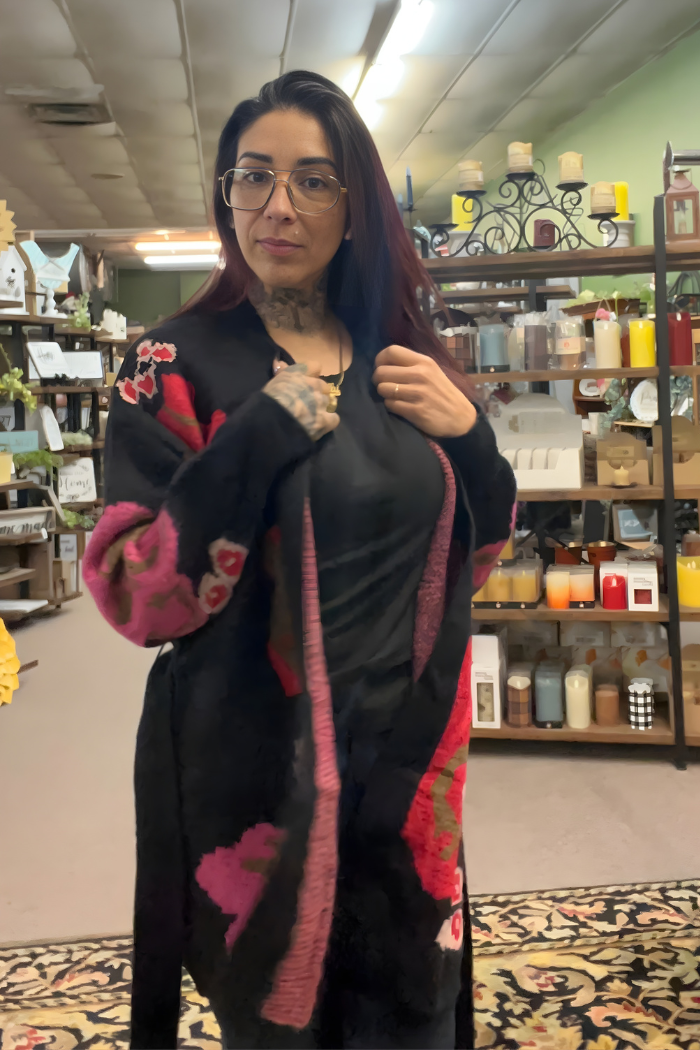 A woman with glasses and tattoos stands indoors in a store filled with candles and decor, wearing the Floral Belted Knit Cardigan featuring cozy pink and red patterns.