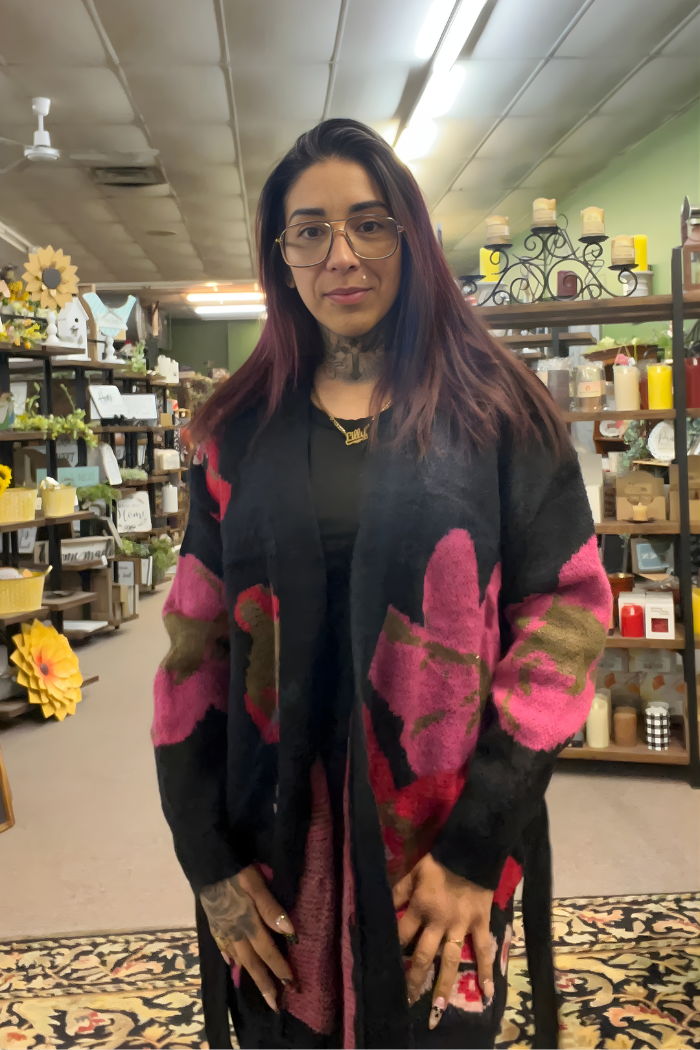 A woman with long dark hair, glasses, and tattoos stands indoors in a shop filled with candles and plants, wearing the Floral Belted Knit Cardigan featuring bright pink and olive floral patterns.