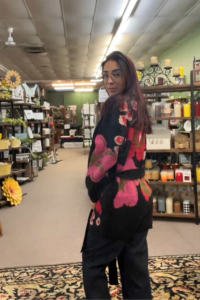 A woman with long hair and glasses wears the Floral Belted Knit Cardigan, featuring black knit fabric with pink and red florals, as she stands in a cozy store with candles and home decor, glancing over her shoulder at the camera.