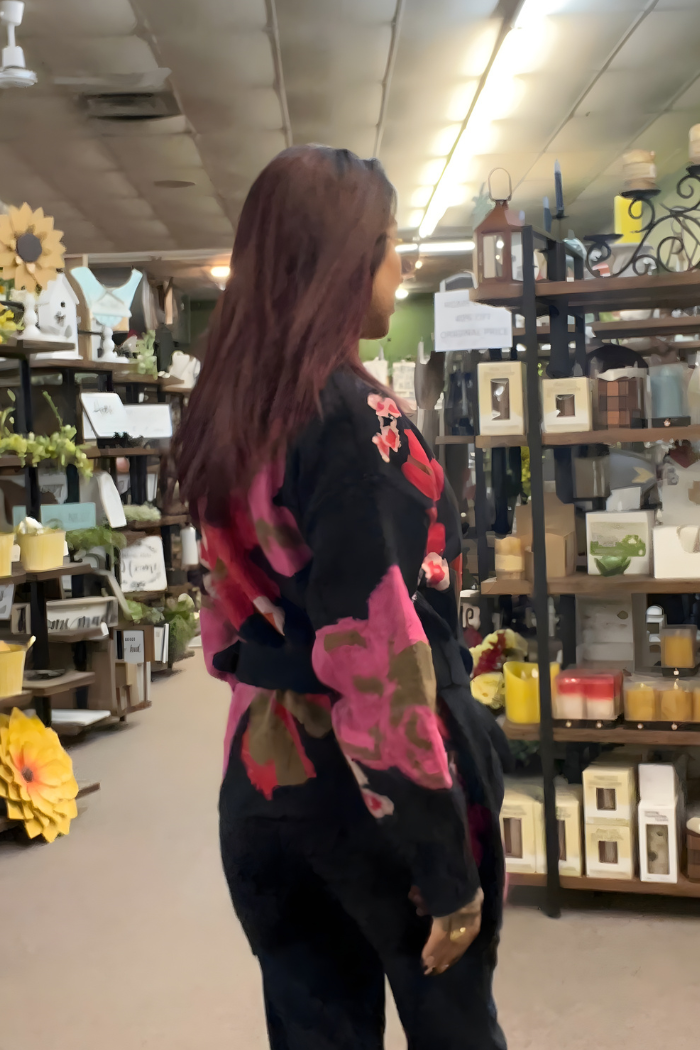 A woman with long, reddish-brown hair wears a Floral Belted Knit Cardigan as she stands in a store aisle lined with shelves of home decor items, candles, and plants.