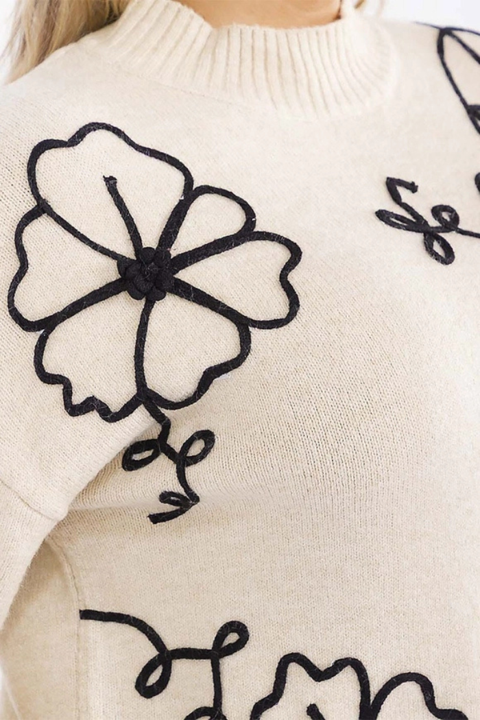 Close-up of the Floral Embroidered Knit Sweater, a cream-colored sweater with black floral designs and swirls on the shoulder and chest. Features a ribbed neckline—perfect for cozy boutique winter sweater collections.