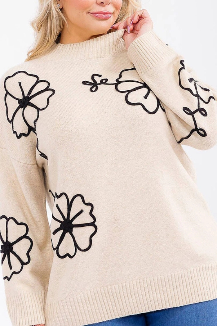 A woman smiles and touches her hair while wearing the Floral Embroidered Knit Sweater, featuring cozy black-outlined floral embroidery, a scalloped neckline hem, and paired with blue jeans.