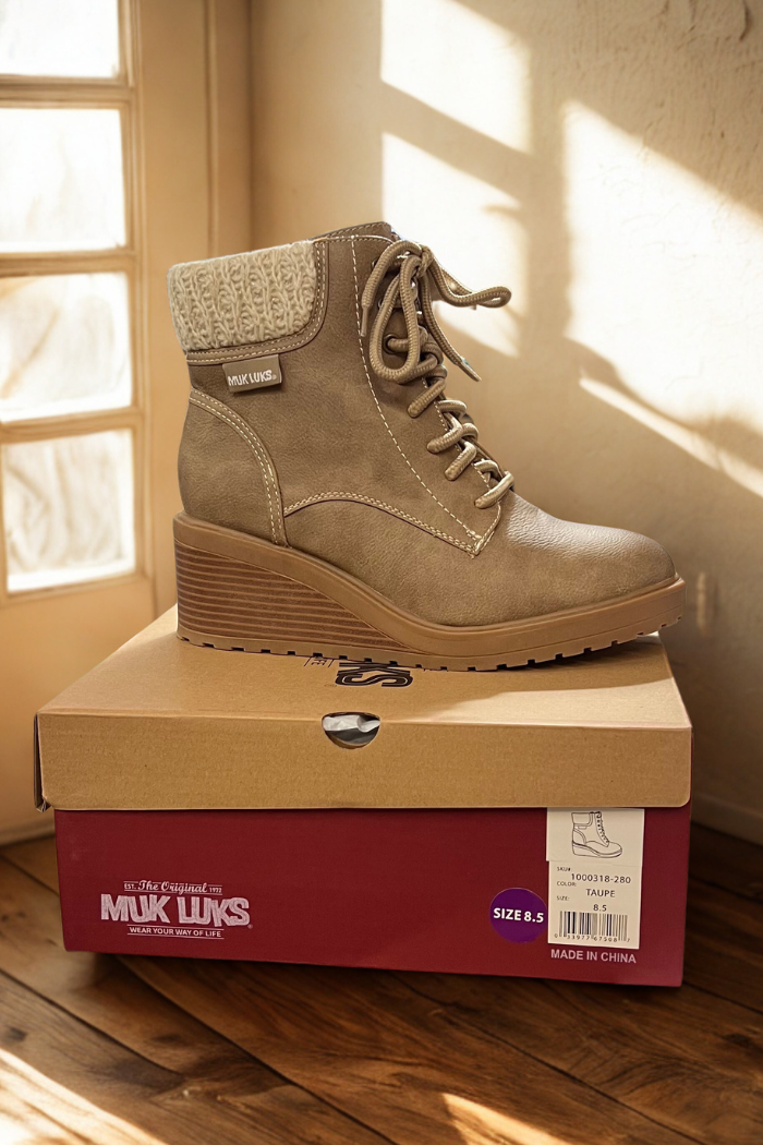 A pair of taupe Sky Solange Wedge Boots with a knit collar sits atop a red MUK LUKS shoe box. Sunlight spills through the window, casting soft shadows on the wooden floor. The box label reads "size 8.5.