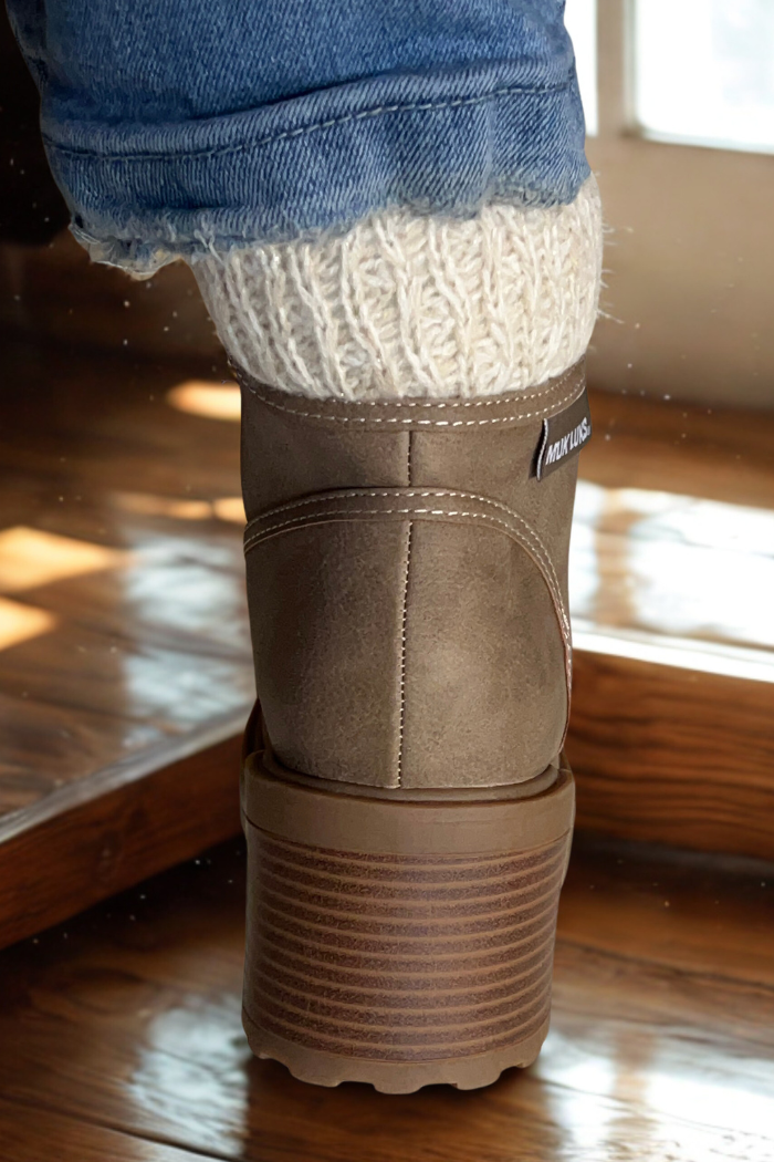 A close-up of someone in cuffed blue jeans, cream knit socks, and taupe Sky Solange Wedge Boots with a ridged sole, standing on a wooden floor by a window.