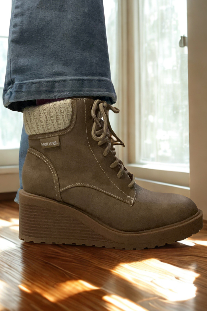 A person wearing blue jeans and Sky Solange Wedge Boots with taupe knit cuffs stands on a sunlit wooden floor near a window.