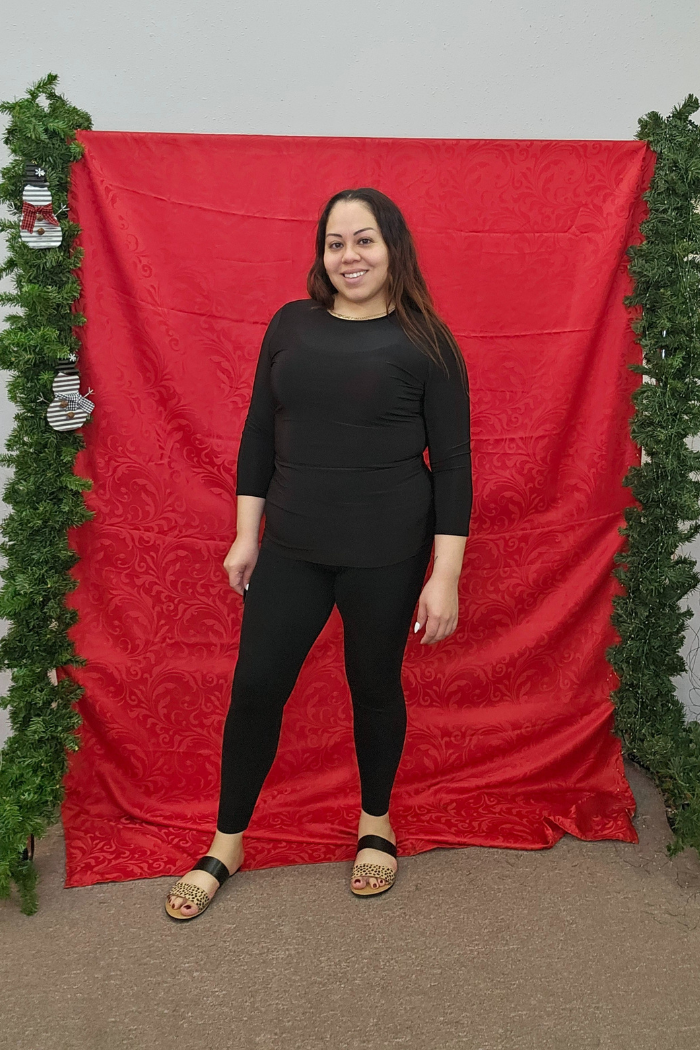 A woman wearing a Wrinkle-Free Layering Top and sandals smiles in front of a red decorative backdrop with garland and holiday stockings at Deja Vu Boutique.