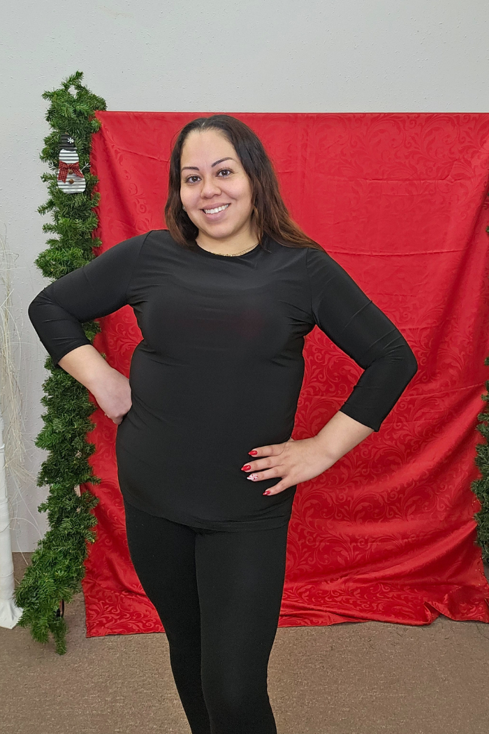 A woman with long brown hair models the Wrinkle-Free Layering Top, paired with black pants. She smiles with one hand on her hip in front of a red backdrop and green garlands.