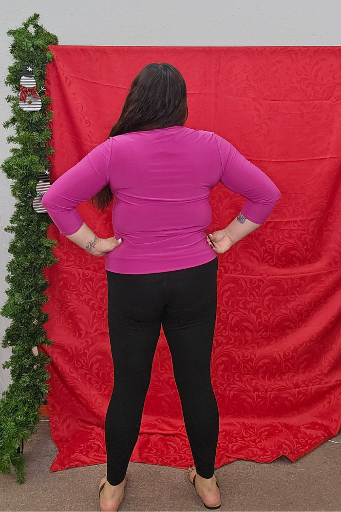 A person with long dark hair, wearing a bright pink Wrinkle-Free Layering Top, black leggings, and sandals stands facing away from the camera in front of a red patterned backdrop with greenery on the left.