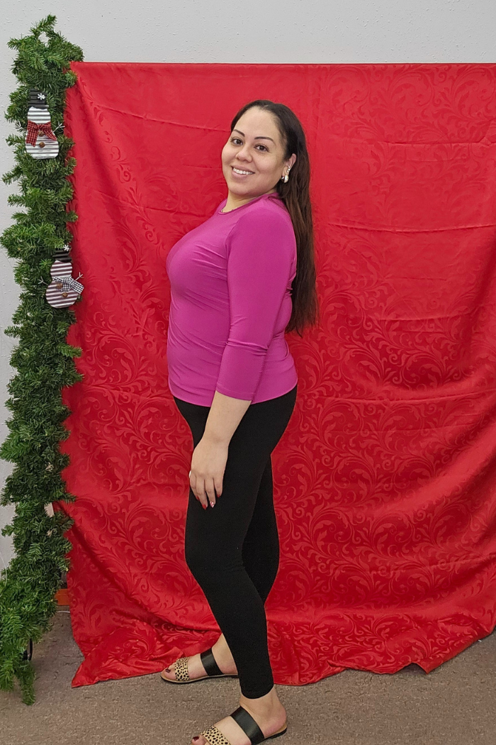 A woman wears the Wrinkle-Free Layering Top in purple and black leggings, standing sideways and smiling at the camera in front of a red backdrop with green garland and stockings, styled by Deja Vu Boutique.