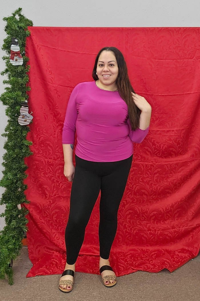 A woman wearing the Wrinkle-Free Layering Top from Deja Vu Boutique in pink, paired with black leggings and sandals, smiles in front of a red fabric backdrop decorated with a green garland on the left.