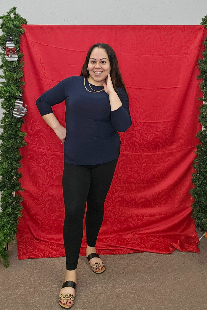 A smiling woman poses before a red patterned backdrop with greenery and small striped mittens, wearing the Wrinkle-Free Layering Top from Deja Vu Boutique, black leggings, and sandals, one hand on her hip and the other touching her face.