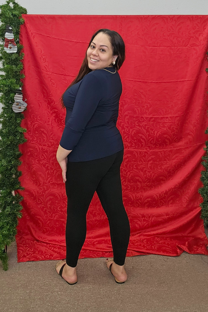 A woman smiles and poses before a red backdrop with green garlands, wearing the Wrinkle-Free Layering Top from Deja Vu Boutique, paired with black leggings and sandals.