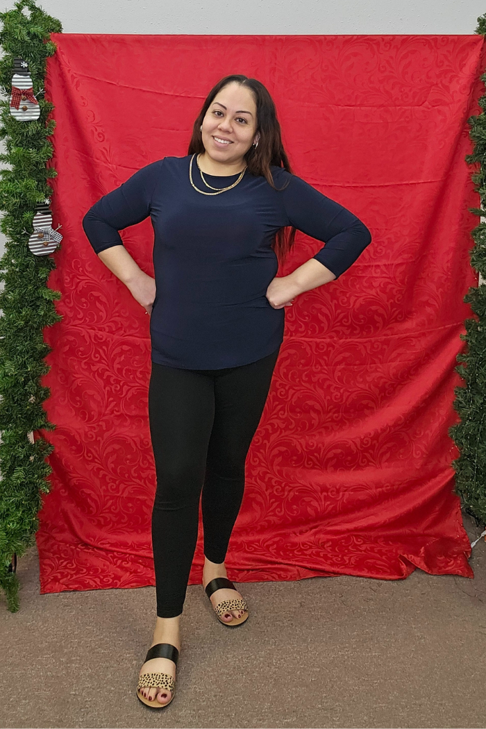 A woman stands smiling before a red decorative backdrop, wearing the Wrinkle-Free Layering Top in navy from Deja Vu Boutique, paired with black leggings and leopard-print sandals. Green garlands with ornaments frame the backdrop.