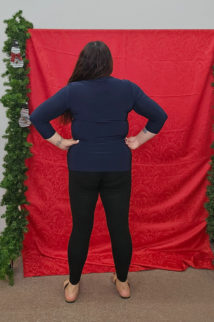 A person with long dark hair stands facing away from the camera, wearing a navy Wrinkle-Free Layering Top, black leggings, and sandals. They’re in front of a red patterned backdrop with greenery on both sides.