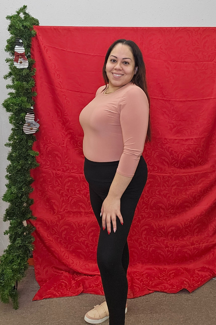 A woman with long dark hair, dressed in a peach Wrinkle-Free Layering Top, black leggings, and beige shoes, smiles in front of a red patterned backdrop beside a garland adorned with small stockings.