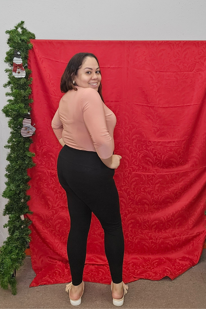 A woman smiles confidently in front of a red decorative backdrop with green garland, wearing the Wrinkle-Free Layering Top from Deja Vu Boutique, black pants, and white shoes, with one hand on her hip.
