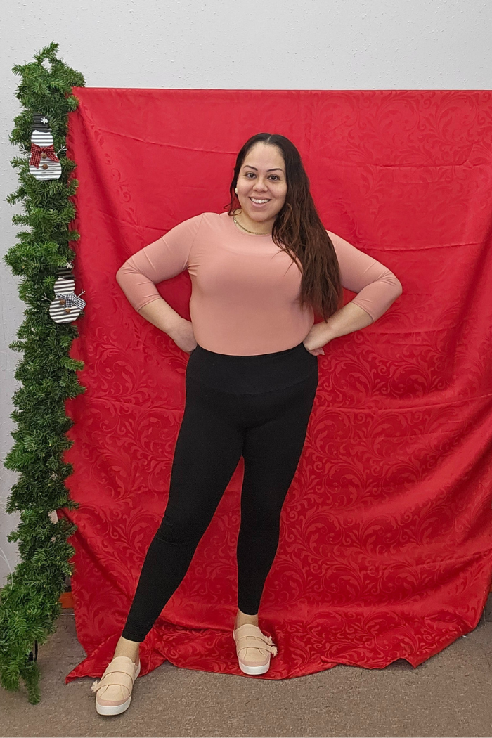 A woman smiles with her hands on her hips in front of a red patterned backdrop. She wears a peach Wrinkle-Free Layering Top from Deja Vu Boutique, black pants, and beige shoes. A vertical garland with silver ornaments hangs at the left.