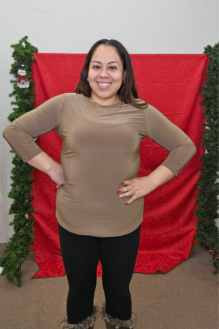 A woman smiles with hands on hips before a red backdrop and green garland, wearing a Wrinkle-Free Layering Top from Deja Vu Boutique, black pants, and fur-trimmed boots.