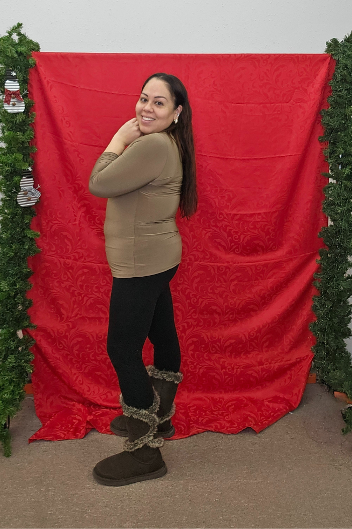 A woman with long brown hair, dressed in a Wrinkle-Free Layering Top from Deja Vu Boutique, black leggings, and brown fur-trimmed boots, smiles while posing before a red patterned fabric backdrop with green garlands.
