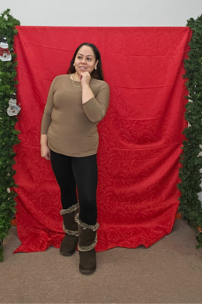 At Deja Vu Boutique, a woman poses before a red patterned backdrop with green garlands, wearing the Wrinkle-Free Layering Top, black leggings, and fur-lined boots. She touches her chin and smiles gently.