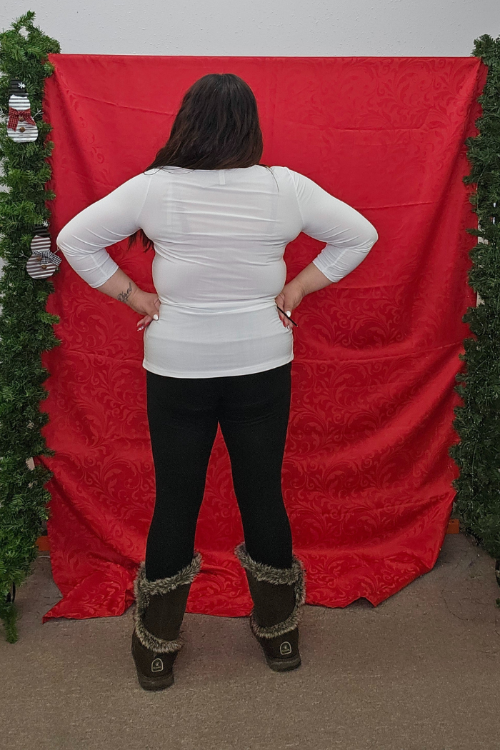 A person with long dark hair faces a red patterned backdrop framed by green garlands, wearing a Wrinkle-Free Layering Top from Deja Vu Boutique, black pants, and fur-lined boots.