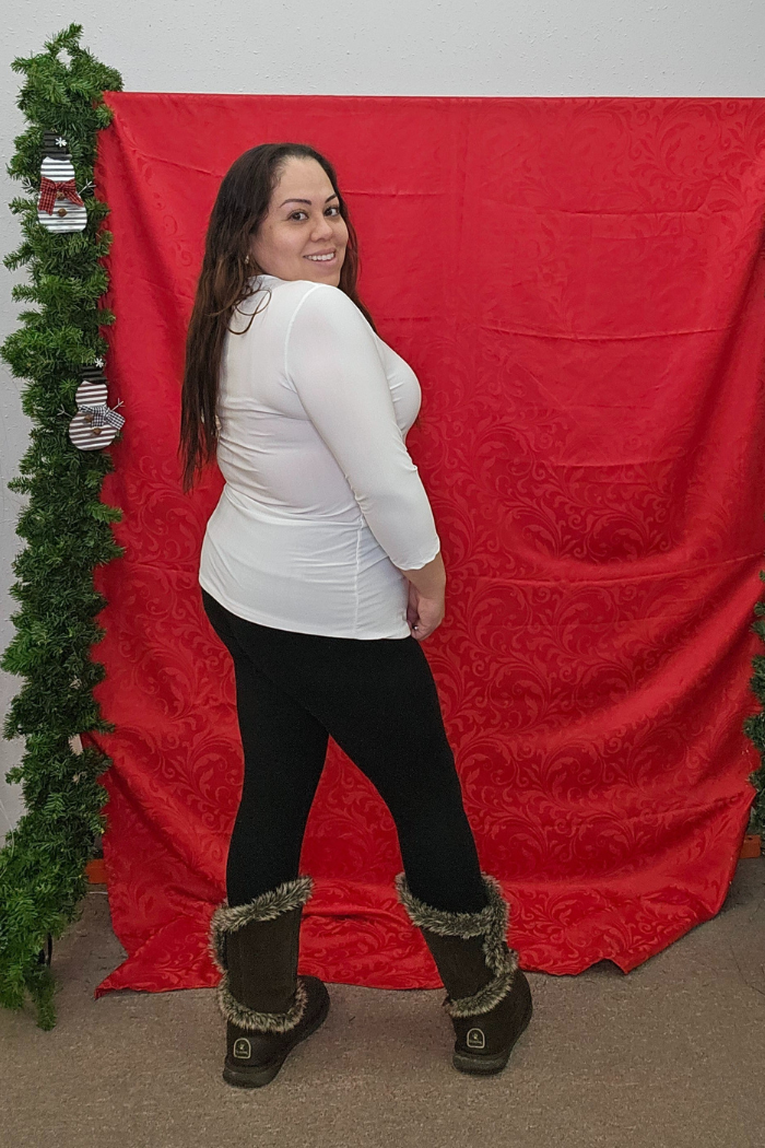 A woman with long dark hair smiles in front of a red decorative backdrop, wearing the Wrinkle-Free Layering Top, black pants, and fur-trimmed boots. Green garlands frame each side of the scene.