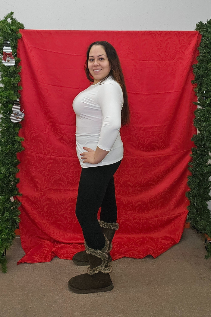 A woman beams at the camera in front of a red backdrop with green garland, wearing a Wrinkle-Free Layering Top, black leggings, and brown fur-trimmed boots. She stands sideways, effortlessly stylish.
