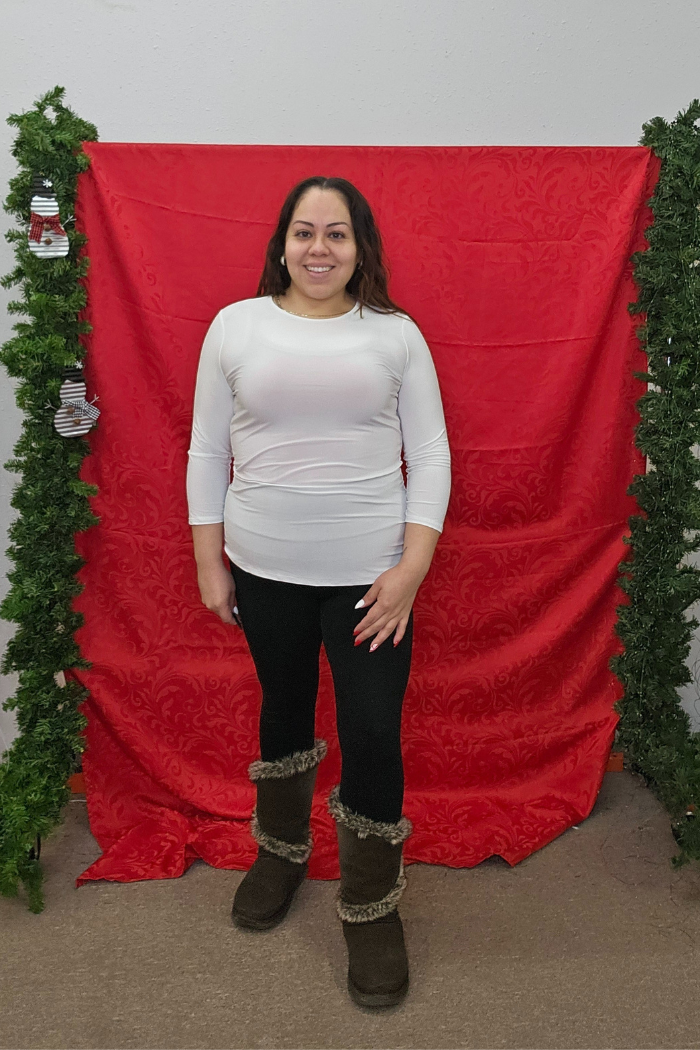 A woman smiles in front of a red fabric backdrop with green garlands. She wears a Wrinkle-Free Layering Top in white, black leggings, and brown furry boots.
