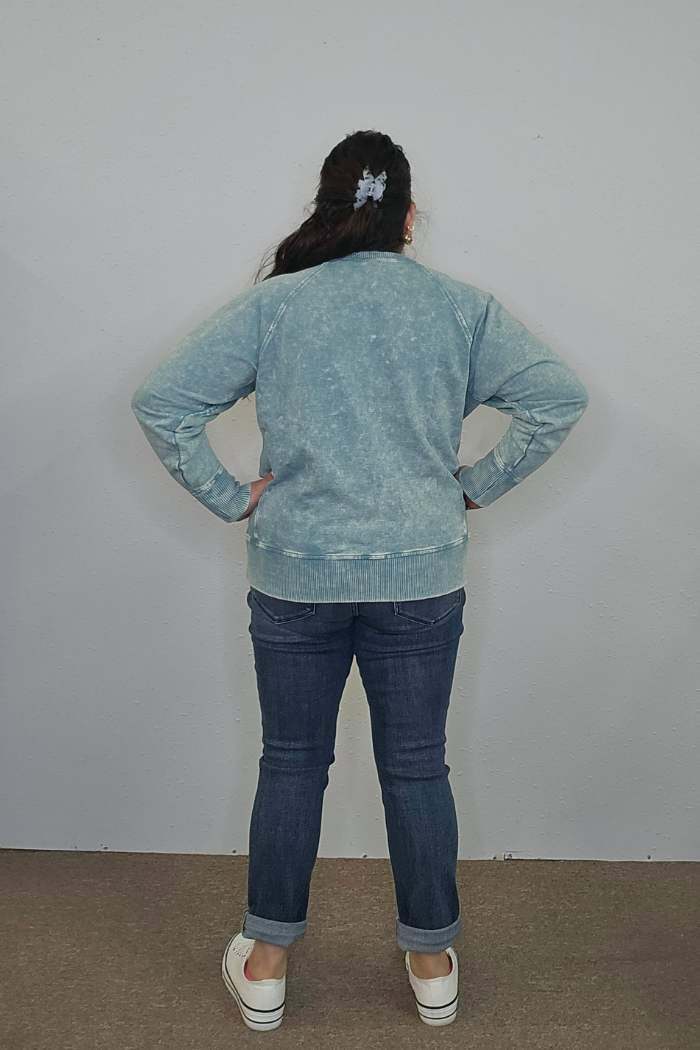 A person with long dark hair in a light blue scrunchie, wearing an oversized Acid Wash Terry Pullover, rolled-up blue jeans, and white slip-on shoes stands facing a plain light-colored wall with hands on hips.