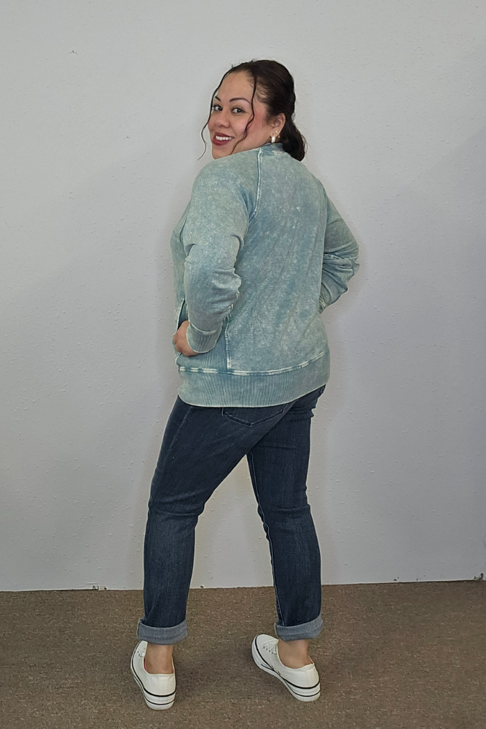 A woman with dark hair in a ponytail smiles while posing in an Acid Wash Terry Pullover, cuffed dark jeans, and white slip-on shoes against a plain, light-colored wall.