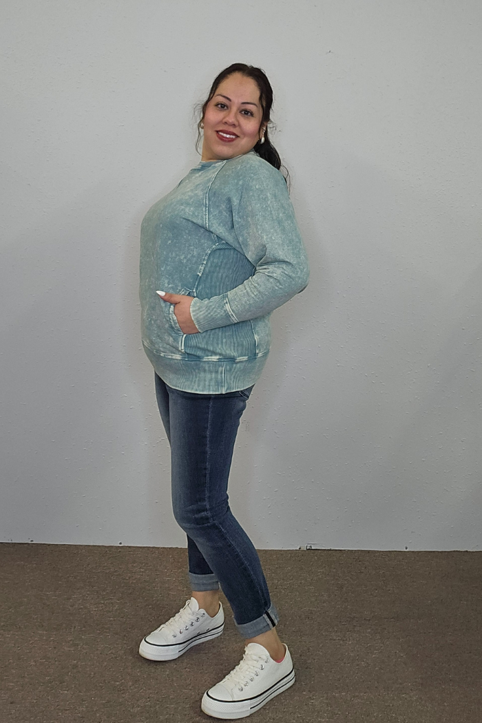 A woman with long dark hair in a ponytail smiles, wearing an Acid Wash Terry Pullover, dark blue cuffed jeans, and white sneakers as she poses against a plain light-colored wall.